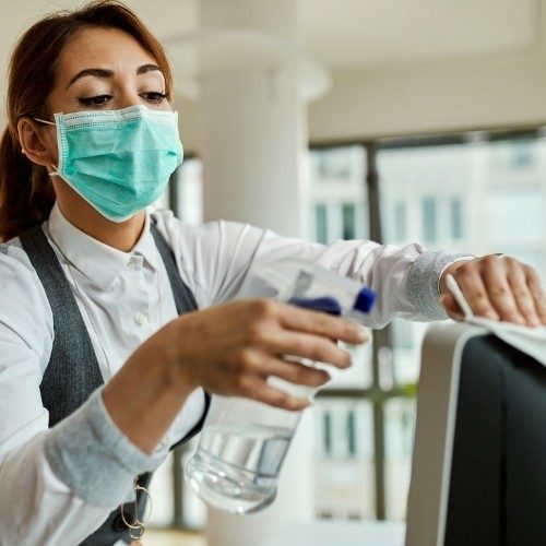 An office cleaning technician wearing a medical face mask and spraying sanitizer onto a desktop monitor.