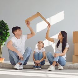 A young mother and father sit surrounding by moving boxes, holding a piece of cardboard above their son's head in the shape of a roof